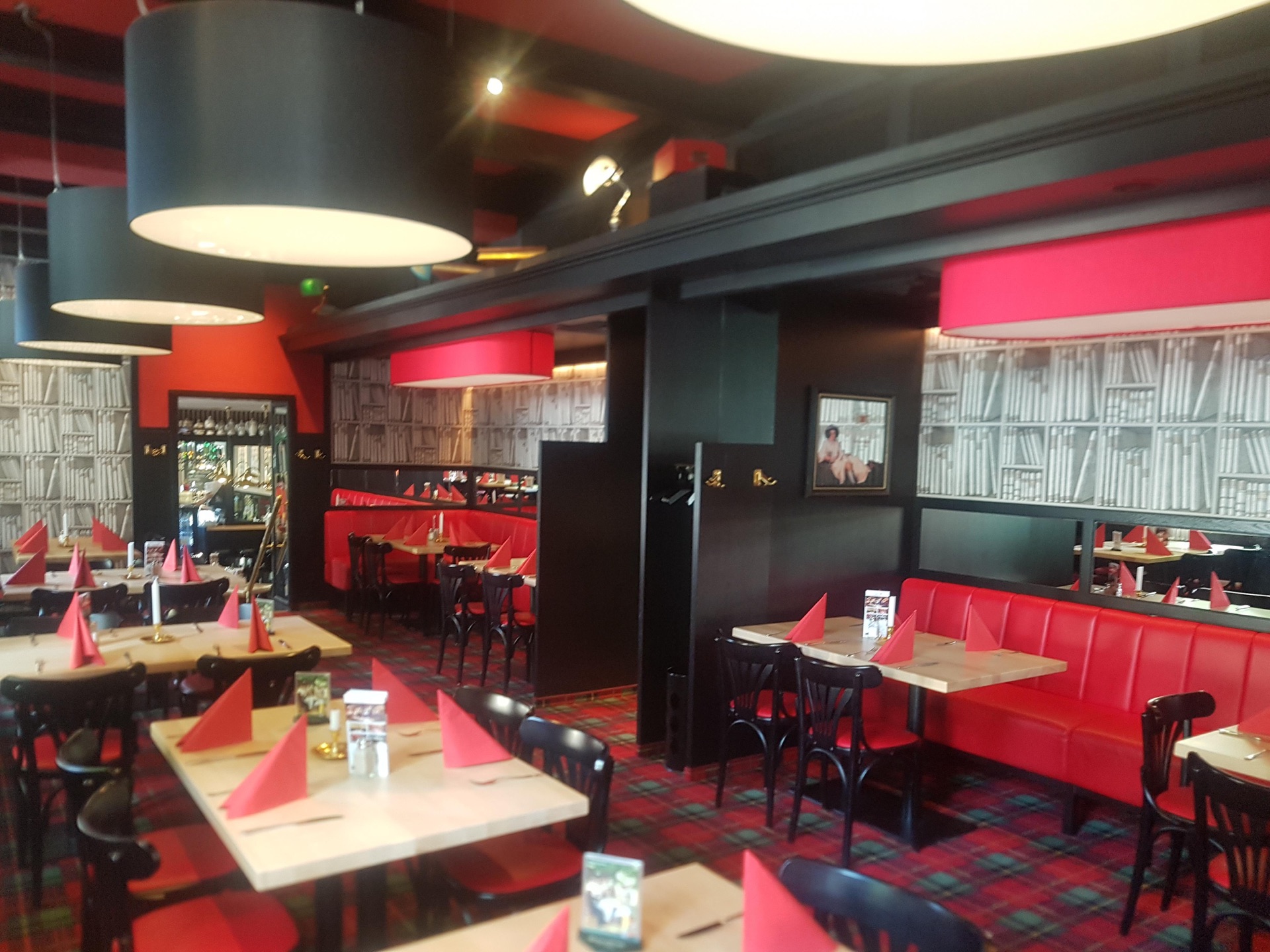 Bild eines Restaurants mit den Hauptfarben Rot und Schwarz. Auffällig sind große Deckenlampen. Auf den Tischen sind rote Servietten. An den Tischen stehen schwarze Stühle und rote Couchs.